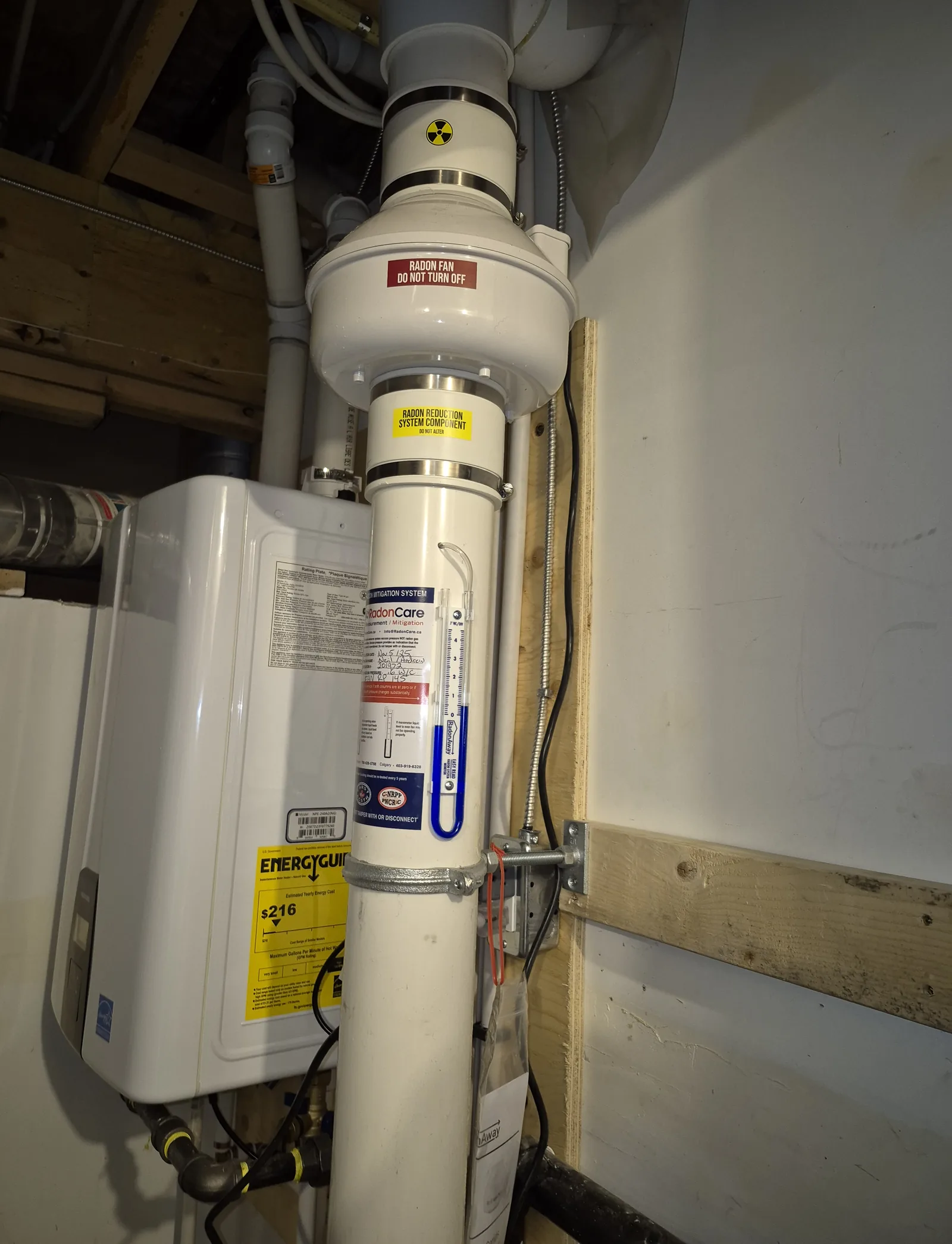 A Radon Fan, Pressure Gauge And Labelled Pipe Beside A Water Heater In An Airdrie Basement