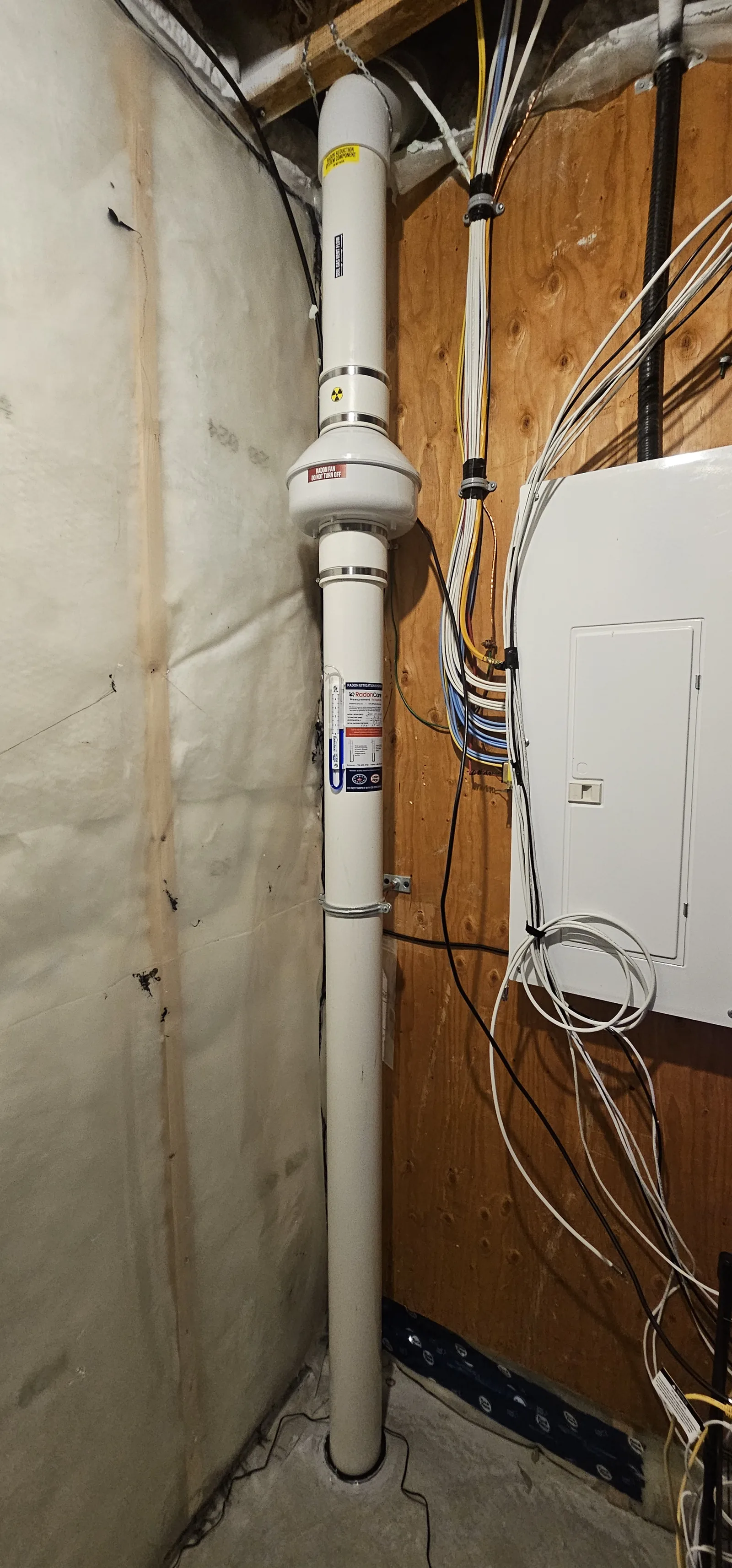 Tall Radon Pipe With The Fan Mounted Beside The Electrical Panel In An Unfinished Cochrane Basement